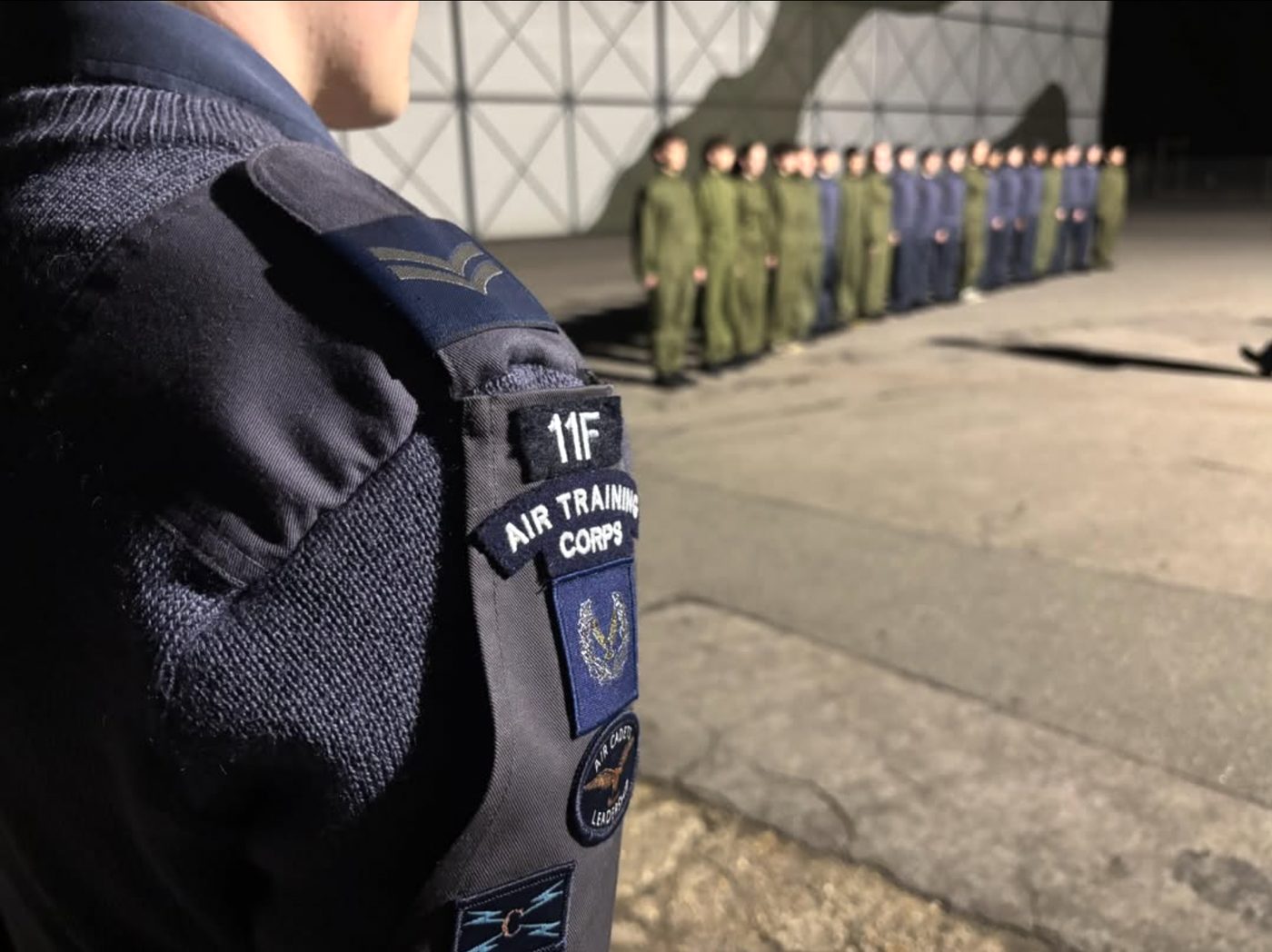 Close-up of a cadet's shoulder, showing 11F and Air Training Corps flashes, with a flight of cadets fallen in behind